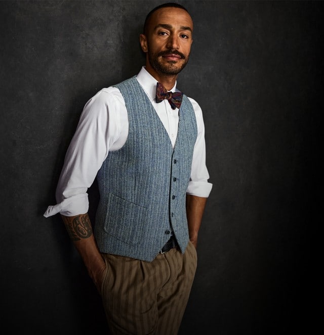 Der Mann trägt ein weißes Hemd mit hochgekrempelten Ärmeln, eine hellblaue Weste und eine beigefarbene Hose sowie eine gemusterte Fliege.