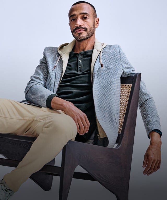 Ein Mann trägt eine hellgraue Jacke über einem dunkelgrünen Henley‑Shirt, kombiniert mit beigefarbenen Hosen und weißen Sneakers. Ein Mann trägt eine hellgraue Jacke über einem dunkelgrünen Henley‑Shirt, kombiniert mit beigefarbenen Hosen und weißen Sneakers.