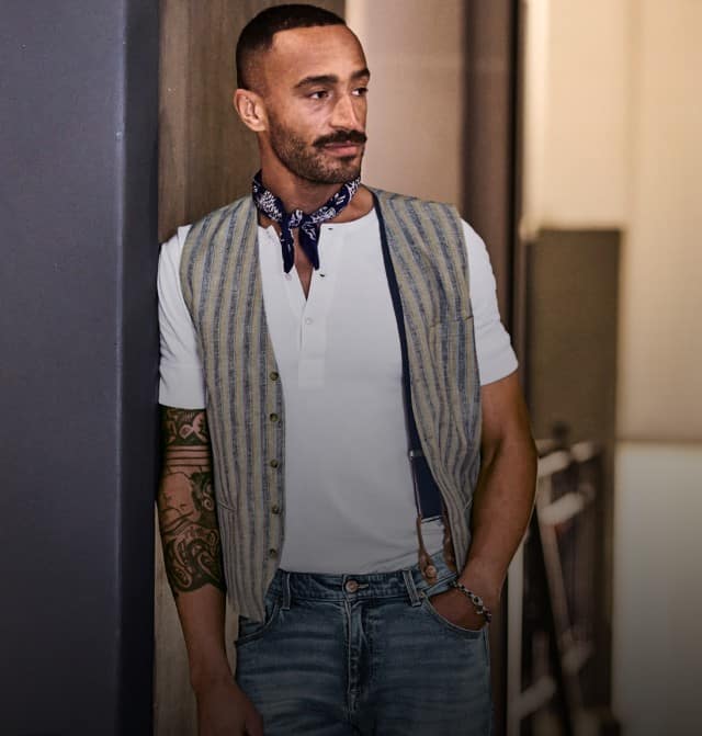 Ein Mann trägt ein weißes Shirt, darüber eine gestreifte Weste und kombiniert dazu eine blaue Jeans sowie ein dunkelblaues Halstuch.