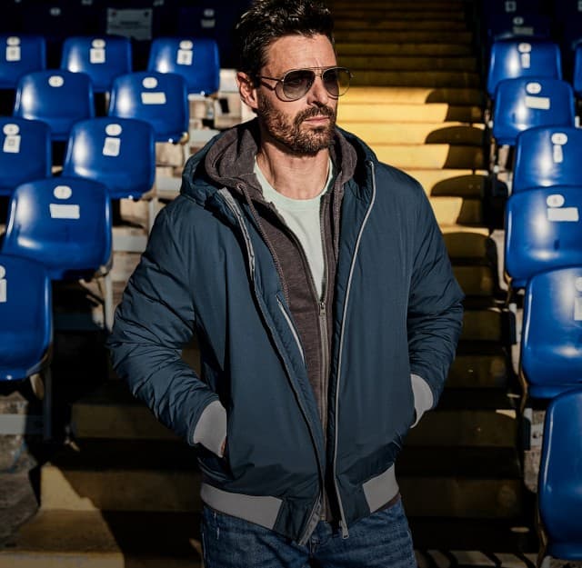Ein Mann trägt eine dunkelblaue Jacke über einem grauen Hoodie und einem hellen T‑Shirt, kombiniert mit einer Jeans und Sonnenbrille.
