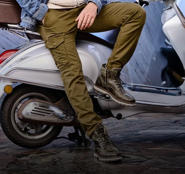 Ein Mann trägt olivgrüne Cargo‑Hosen und robuste braune Lederboots, während er auf einem weißen Motorroller sitzt.