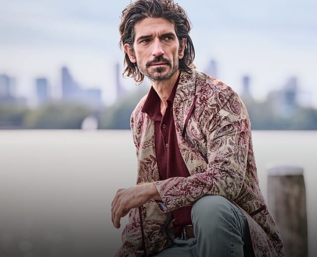 Ein Mann trägt ein burgunderrotes Shirt unter einem langen, beigefarbenen Mantel mit bordeauxrotem Paisley‑Muster, kombiniert mit einer hellen Hose.