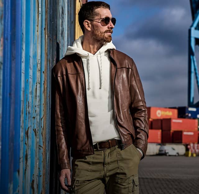Ein Mann trägt eine braune Lederjacke über einem cremefarbenen Hoodie und kombiniert dazu olivgrüne Cargo‑Hosen.
