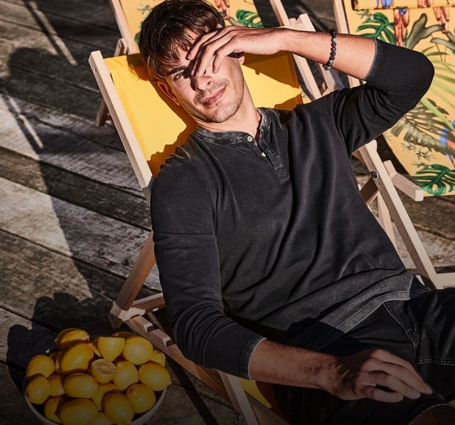 Ein Mann trägt ein dunkelgraues Henley‑Shirt mit langen Ärmeln und eine schwarze Shorts, während er entspannt in einem gelben Liegestuhl sitzt.