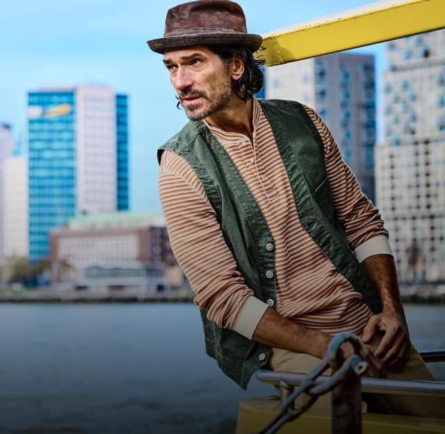 Ein Mann mit rot gestreiftem Longsleeve und einer grünen Weste steht auf einem Boot, hinter ihm ist die Skyline einer Stadt zu sehen.