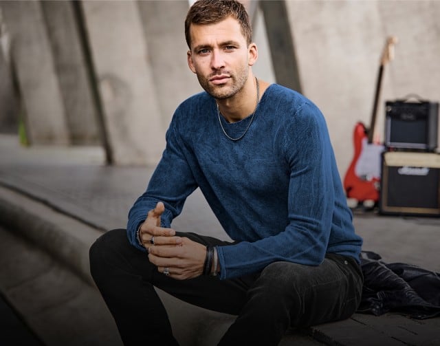 Ein Mann bekleidet mit einem blauen Pullover und einer dunkelgrauen Jeans sitzt entspannt auf einer Betonstufe.