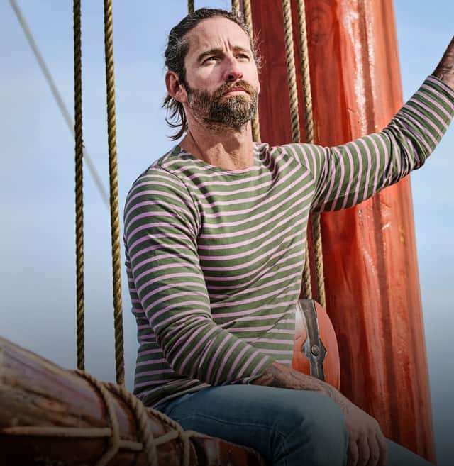 Ein Mann trägt ein gestreiftes Langarmshirt und eine hellblaue Jeans uns sitzt auf einem Segelboot.