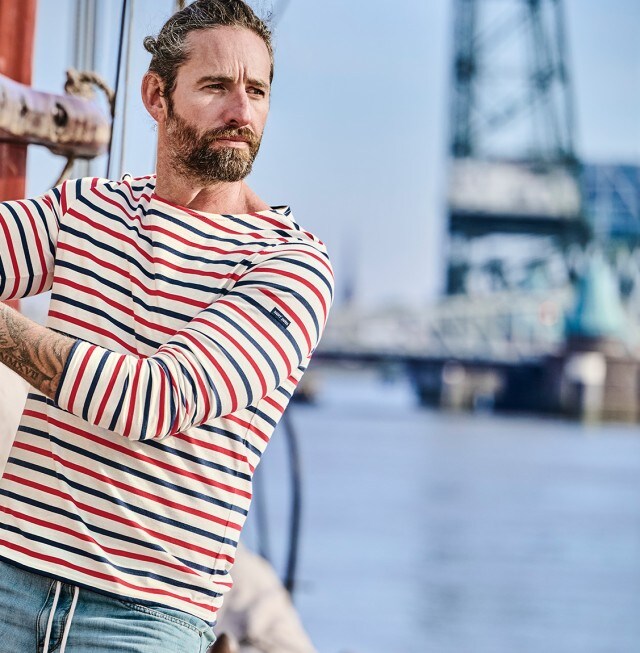 Ein Mann trägt ein gestreiftes Langarmshirt und steht auf einem Segelboot