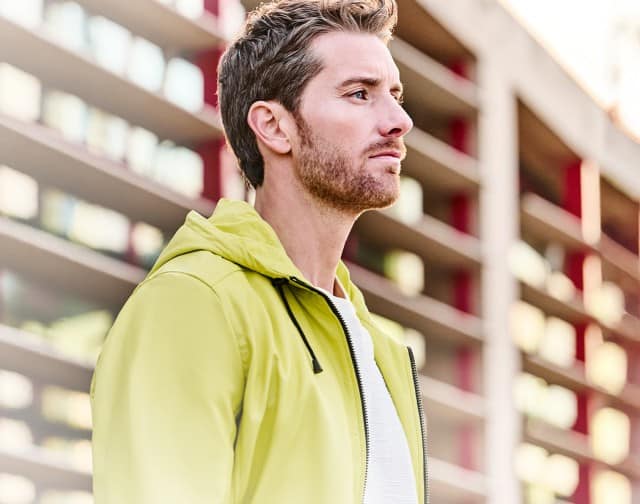 Ein Mann bekleidet mit einer gelben Jacke und einem weißen Shirt, steht vor einer Fensterfassade und blickt in die Ferne