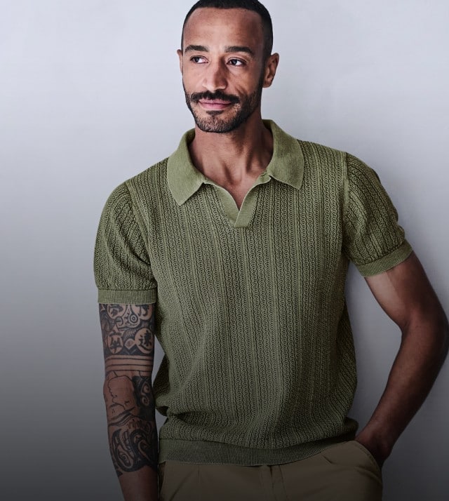 Auf dem Bild ist ein Mann zu sehen der mit einem grünen Strickpolo bekleidet vor einer grauen Wand steht.