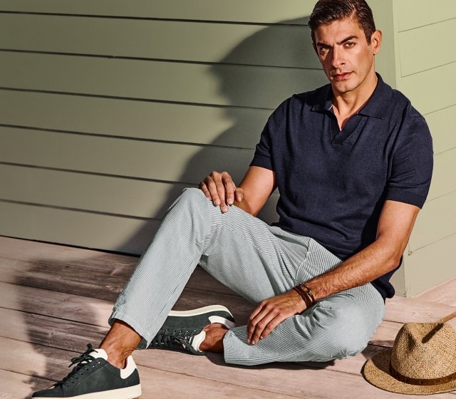 Ein Mann bekleidet mit einem dunklen Polo und einer gestreiften Sommerhose sitzt entspannt und lehnt an einer Hauswand