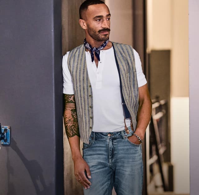 Ein Mann trägt ein weißes T-Shirt in Kombination mit einer gestreiften Weste und einer hellen Jeans, er lehnt sich an eine graue Wand.