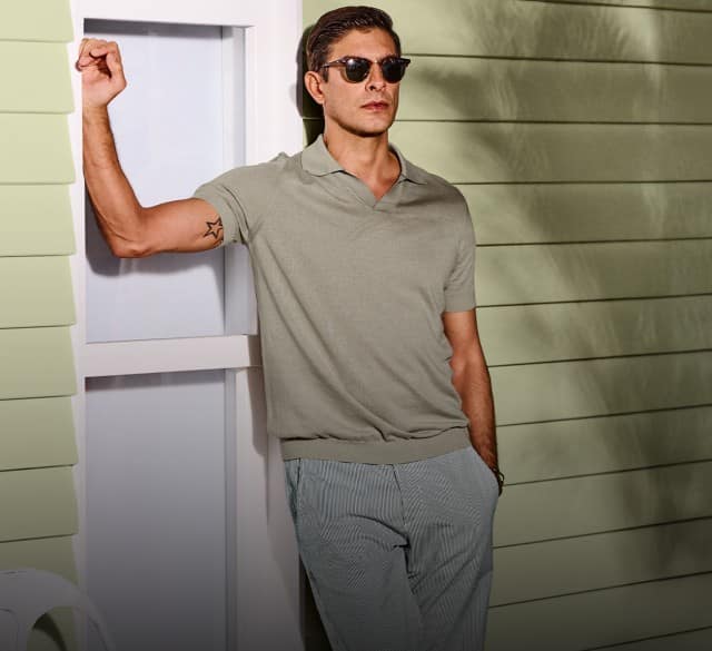 Ein Mann bekleidet mit einem beigen PoloShirt steht angelehnt an einem Beachouse.