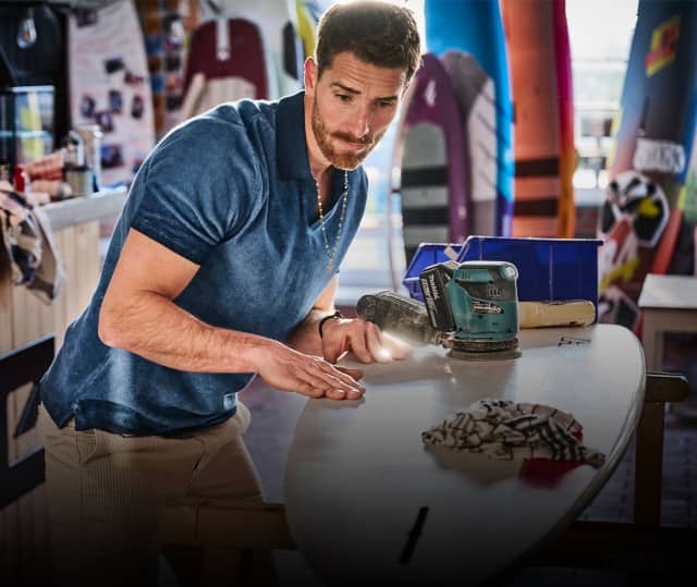 Ein Mann bekleidet mit einem blauen Polo und einer beigefarbenen Chino steht in einem Laden und poliert ein Surfbrett.