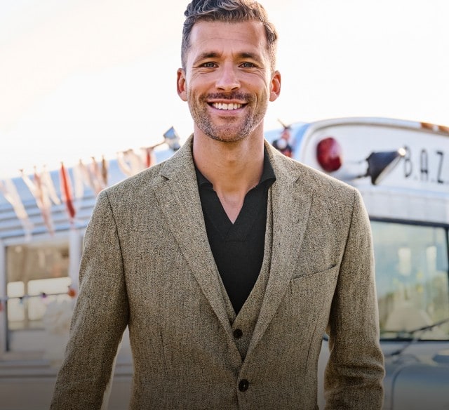 Ein Mann steht gut gelaunt vor einem Partybus. Bekleidet ist er mit einem grauen Anzug und darunter trägt er ein schwarzes Hemd.