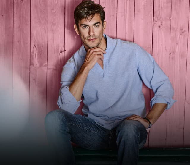 Ein Mann bekleidet mit einem hellblauen Leinenhemd sitzt angelehnt an eine Holzwand und schaut in die Kamera.