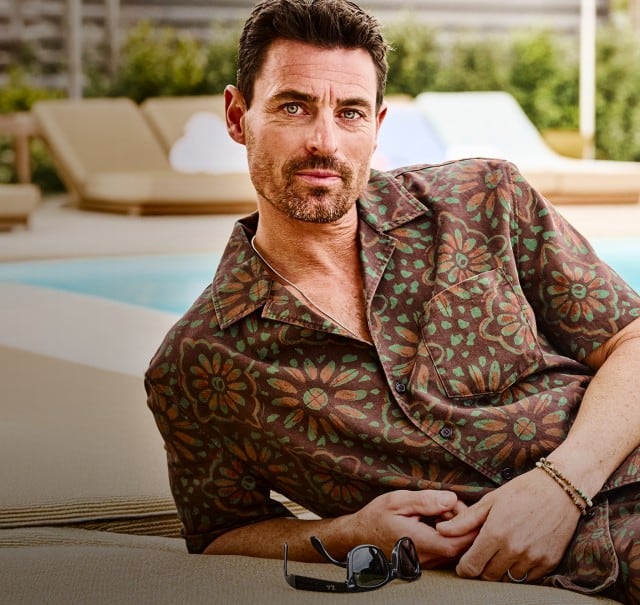 Zu sehen ist eine Person, die auf einer Liege am Rand eines Pools liegt, legere gemusterte Kleidung trägt, während im Hintergrund weitere Liegen und eine begrünte Außenanlage erkennbar sind.