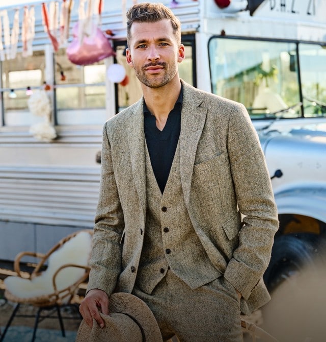 Zu sehen ist eine Person in einem hellen, dreiteiligen Anzug, die im Freien vor einem dekorierten, retro‑artigen Bus steht, wobei im Hintergrund eine ländliche Umgebung mit Grünflächen und bunten Hängedekorationen erkennbar ist.