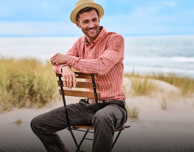 Zu sehen ist eine Person mit Strohhut und gestreiftem Hemd, die auf einem Klappstuhl in sandigen Dünen sitzt, während im Hintergrund das Meer und ein heller Himmel zu erkennen sind.
