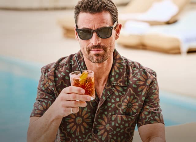 Ein Mann mit einem bedruckten Hemd sitzt mit einem Drink in der Hand vor einem Pool.