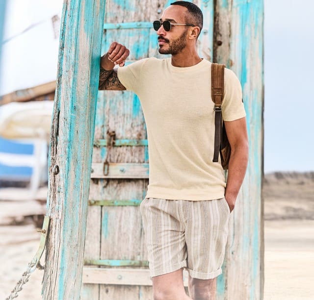 Ein Mann steht im Strandoutfit bestehend aus Shorts und T-Shirt am Strand und lehnt an einer blauen Holzwand.