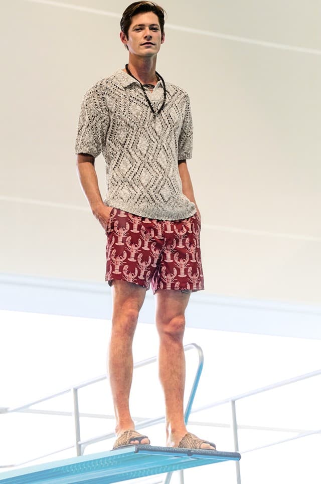 Ein Mann trägt ein helles Strickhemd und rote Shorts mit Muster und steht auf einem Sprungbrett