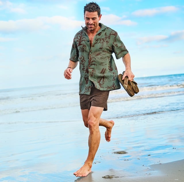 Ein Mann bekleidet mit einem Hemd und einer Shorts läuft entspannt am Strand entlang