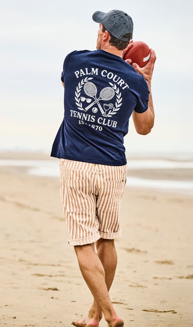 Rückansicht von einem Mann bekleidet mit einem Print Shirt und einer gestreiften Hose steht am Strand und hält einen Baseball in der Hand.