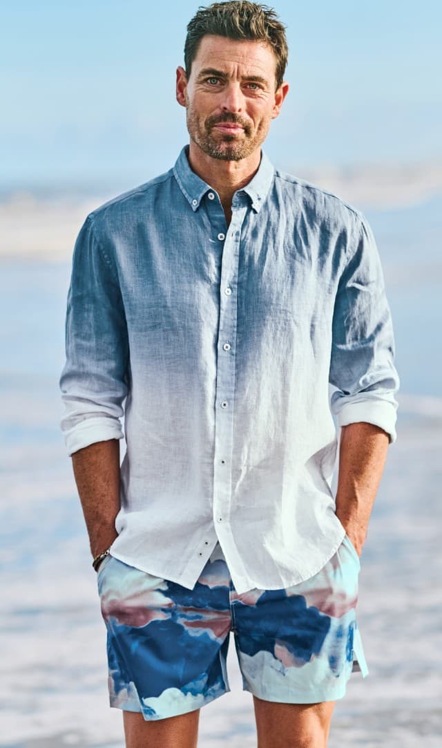 Ein Mann steht mit einem blauen langärmeligen Hemd und einer Shorts bekleidet am Strand, hinter ihm ist das Meer zu sehen.