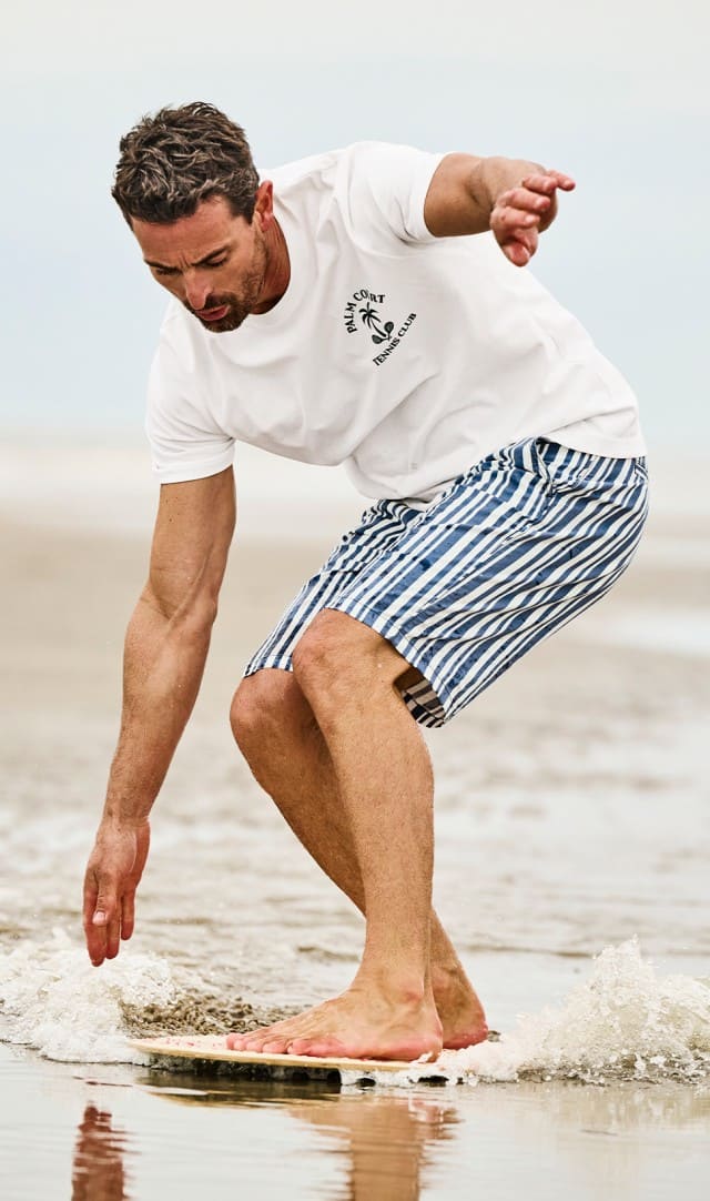 Ein Mann bekleidet mit einem weißen Shirt und einer gestreiften Shorts steht auf einem Surfbrett am Strand, 