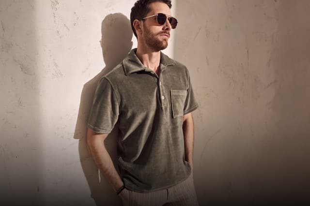 Auf dem Bild steht eine Person mit Sonnenbrille und grünem Poloshirt mit Brusttasche vor einer hellen, strukturierten Wand, wobei Licht und Schatten auf den Hintergrund fallen.