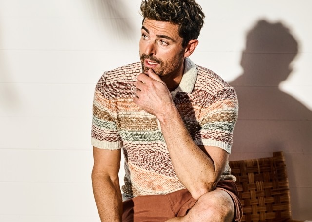 Auf dem Bild ist ein Mann zu sehen, der ein bunt gemustertes Strick-Poloshirt und braune Shorts trägt.