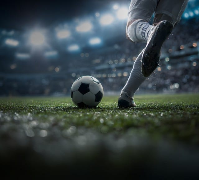 Ein Fußballspieler in weißer Sportkleidung trifft auf einem beleuchteten Stadionrasen einen Ball, während im Hintergrund ein ausverkauftes Stadion mit Flutlicht zu sehen ist.