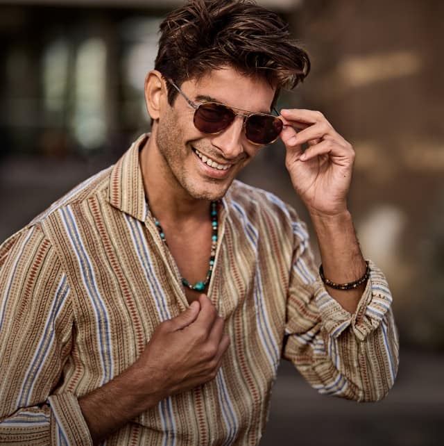 Auf dem Bild ist eine Person in einem gestreiften Hemd zu sehen, die lächelnd eine Sonnenbrille anhebt und dabei im Freien vor einem verschwommenen Hintergrund steht.