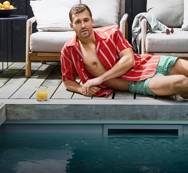 Auf dem Bild liegt eine Person in einem geöffneten rot gestreiften Hemd und grünen Shorts entspannt neben einem Pool auf einer Holzterrasse, vor Outdoor-Möbeln und einem Getränk auf dem Boden. Auf dem Bild liegt eine Person in einem geöffneten rot gestreiften Hemd und grünen Shorts entspannt neben einem Pool auf einer Holzterrasse, vor Outdoor-Möbeln und einem Getränk auf dem Boden.