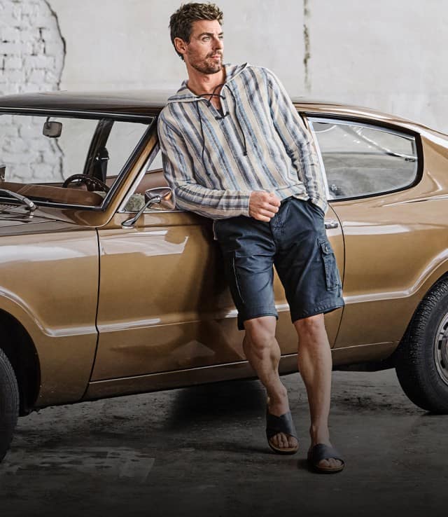 Auf dem Bild lehnt eine Person in einem gestreiften Hemd und Shorts an einem goldfarbenen Oldtimer-Auto in einer Garage oder Halle mit Backsteinwand.