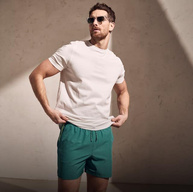 Auf dem Bild steht eine Person mit Sonnenbrille, weißem T‑Shirt und grünen Shorts vor einer schlichten Wand im Licht-Schatten-Spiel und hält den Saum des Shirts mit beiden Händen.