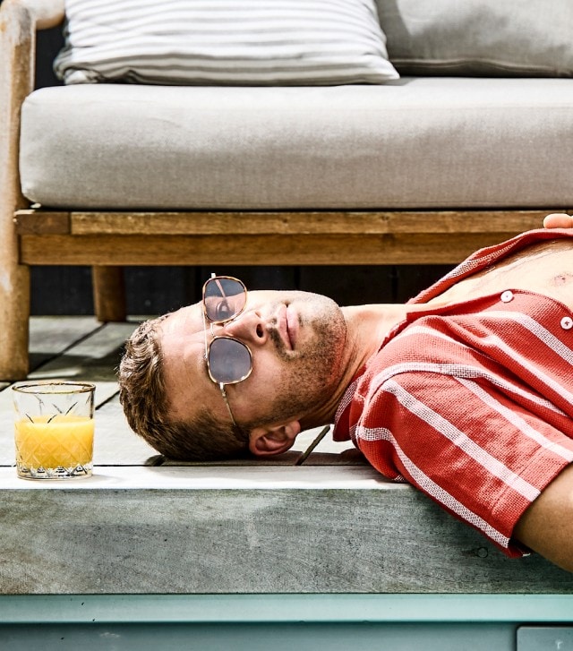 Auf dem Bild liegt ein Mann entspannt in Badehose und offenem Hemd am Rand eines Pools, trägt Sonnenbrille und hat ein Glas Orangensaft neben sich stehen.