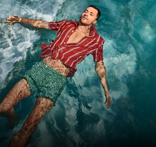 Auf dem Bild ist ein Mann zu sehen, der entspannt auf dem Rücken im Wasser treibt und dabei eine grün gemusterte Badehose sowie ein rot-weiß gestreiftes, offenes Hemd trägt.