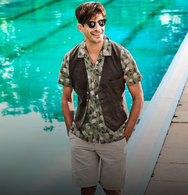Eine Person in sommerlicher Kleidung mit gemustertem Hemd, Weste, Shorts und Sonnenbrille steht locker an einem türkisfarbenen Poolrand auf einem gepflasterten Weg.