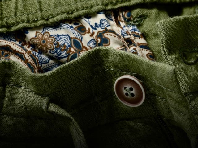 Eine detaillierte Nahaufnahme zeigt den Bund einer grünen Shorts mit Knopf und gemustertem Innenfutter, während rechts daneben die gleiche Shorts freigestellt vor hellem Hintergrund dargestellt ist.