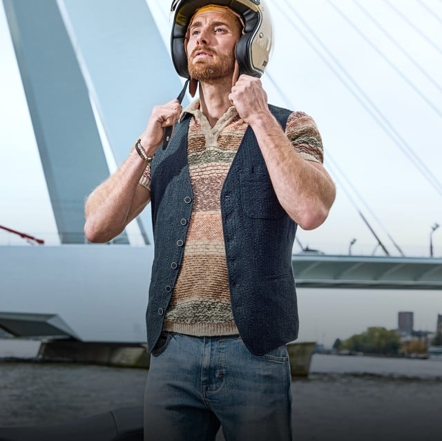 Auf dem Bild steht eine Person neben einem schwarzen Motorroller am Wasser vor einer modernen Brücke und setzt sich einen Helm auf.