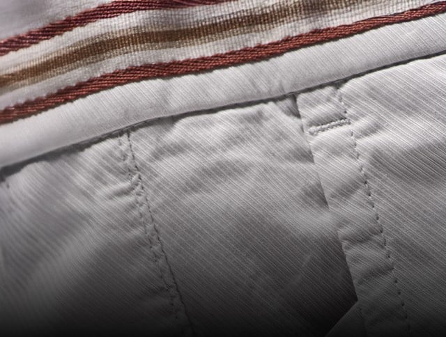 Auf dem Bild sind beige kurze Hosen (Shorts) zu sehen, die aus einem fein strukturierten Stoff bestehen und oben einen Bund mit dezenten rot-braunen Streifen als Detail haben.