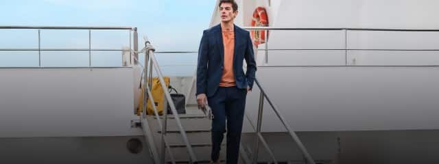 Auf dem Bild trägt die Person einen dunkelblauen Anzug mit passender Hose, darunter ein einfarbiges T‑Shirt in einem warmen Orange‑Ton sowie helle Sneaker mit farbigen Details.