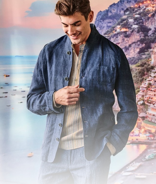Auf dem Bild ist ein junger Mann zu sehen, der lächelnd in einem eleganten Outfit mit blauem Sakko vor einer malerischen Kulisse aus Küste und farbenfrohen Häusern steht.