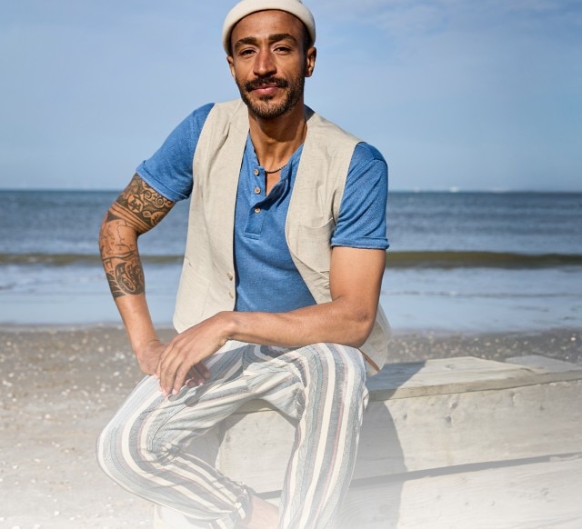 Auf dem Bild sitzt ein Mann der  eine helle Mütze, ein blaues T-Shirt, eine beigefarbene Weste und eine gestreifte, helle Hose trägt.
