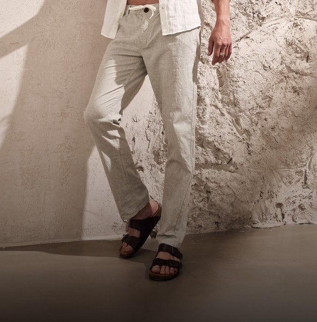 Zu sehen ist der untere Teil einer Person in heller Leinenkleidung und dunklen Sandalen, die vor einer hellen, strukturierten Wand steht, während ein langer Schatten diagonal über die Szene fällt.