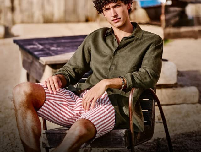 Eine Person in einem olivgrünen Leinenhemd und rot-weiß gestreiften Shorts sitzt entspannt auf einem Stuhl in einer sonnigen Dünenlandschaft mit Sand und Küstenpflanzen.