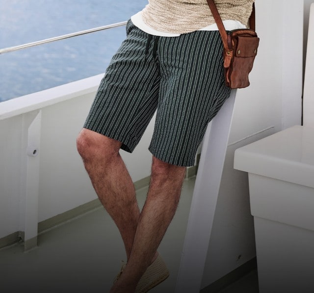 Auf dem Bild ist eine Person zu sehen, die eine gestreifte Shorts, ein beiges Oberteil und eine kleine braune Umhängetasche trägt.