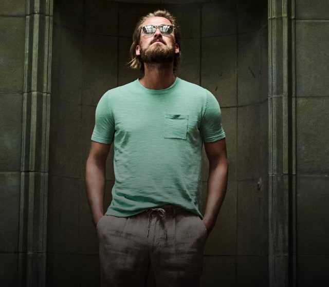 Auf dem Bild ist ein Mann mit Sonnenbrille zu sehen, der ein grünes T-Shirt mit Brusttasche und eine beige Leinenhose trägt.